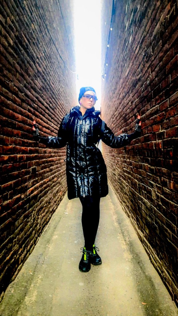 Kae Audhild between buildings, fitness model backlit in alleyway, wearing shein black shiny puffer bomber jacket with matching long hooded vest, ouija beanie, big luxury look sunglasses, Prozis jet high waist leggings, puma twitch trail runners sneakers, grey and black striped knit fingerless half finger gloves, compression gloves, winter Cyber goth alt girl fashion, shimmery pink ethereal mystical faery fairy makeup, holographic highlighters, December 2023 in Owego ny