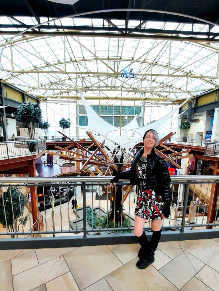 Kae Audhild at Destiny USA mall in Syracuse, NY, Central New York attractions, alternative goth punk lifestyle fashion beauty travel blogger, keracolor silver blue toning semipermanent hair toning color conditioner, killstar x rob zombie official print bardeau style goth punk skater dress, shein black faux leather studded buckle corset style waist belt, shein sxy shiny black puffer bomber jacket, black knee high socks from Amazon, black vegan leather Demonia Lilith-211 mid-calf buckle side zip boots, best of winter 2023 goth fashion, tattooed pierced alt models, thick coarse curly bleached icy blue hair, real natural scandi curls waves, women's afab nonbinary asymmetrical undercut hairstyles