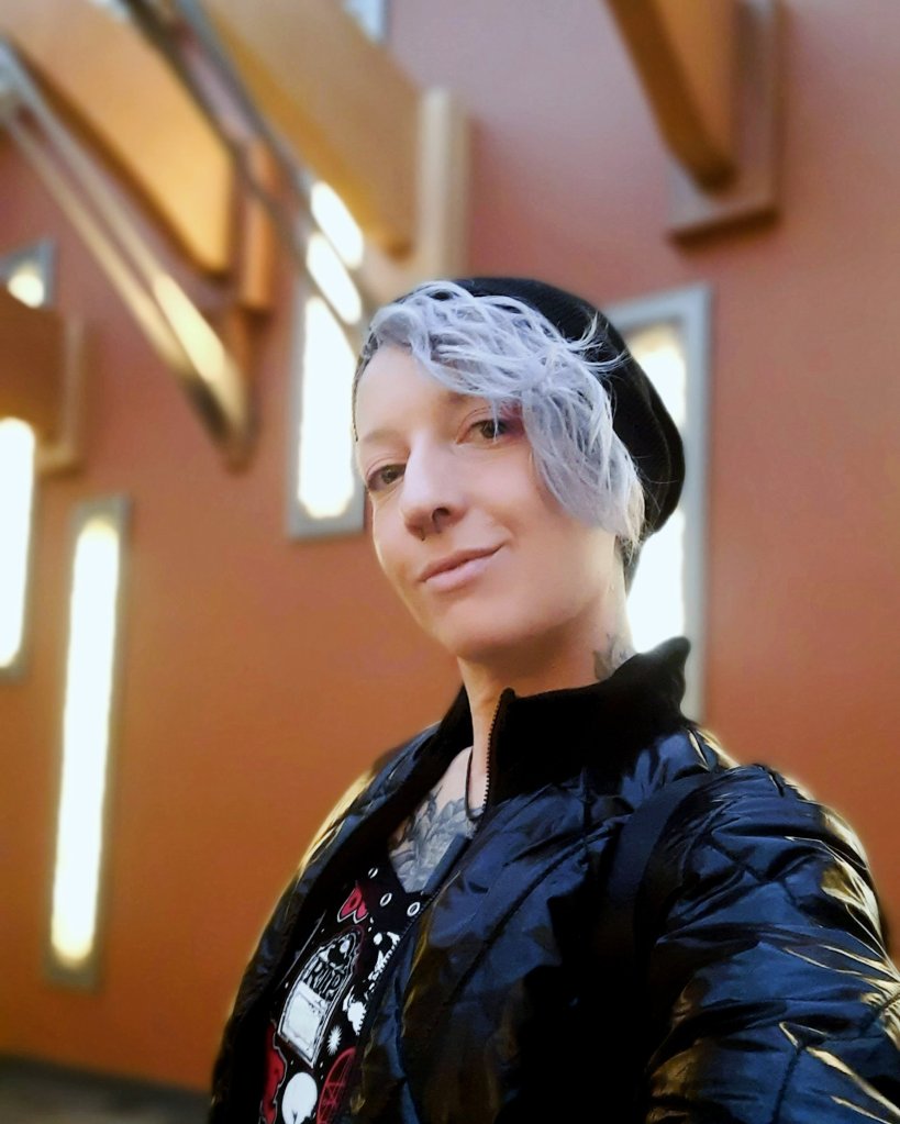 Kae Audhild at Destiny USA mall in Syracuse, NY, Central New York attractions, alternative goth punk lifestyle fashion beauty travel blogger, keracolor silver blue toning semipermanent hair toning color conditioner, killstar x rob zombie official print bardeau style goth punk skater dress, shein black faux leather studded buckle corset style waist belt, shein sxy shiny black puffer bomber jacket, black knee high socks from Amazon, black vegan leather Demonia Lilith-211 mid-calf buckle side zip boots, best of winter 2023 goth fashion, tattooed pierced alt models, thick coarse curly bleached icy blue hair, real natural scandi curls waves, women's afab nonbinary asymmetrical undercut hairstyles