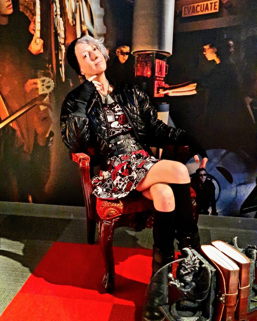 Kae Audhild at 5 Wits adventures at Destiny USA mall in Syracuse, NY, Central New York attractions, alternative goth punk lifestyle fashion beauty travel blogger, keracolor silver blue toning semipermanent hair toning color conditioner, killstar x rob zombie official print bardeau style goth punk skater dress, shein black faux leather studded buckle corset style waist belt, shein sxy shiny black puffer bomber jacket, black knee high socks from Amazon, black vegan leather Demonia Lilith-211 mid-calf buckle side zip boots, best of winter 2023 goth fashion, tattooed pierced alt models, thick coarse curly bleached icy blue hair, real natural scandi curls waves, women's afab nonbinary asymmetrical undercut hairstyles