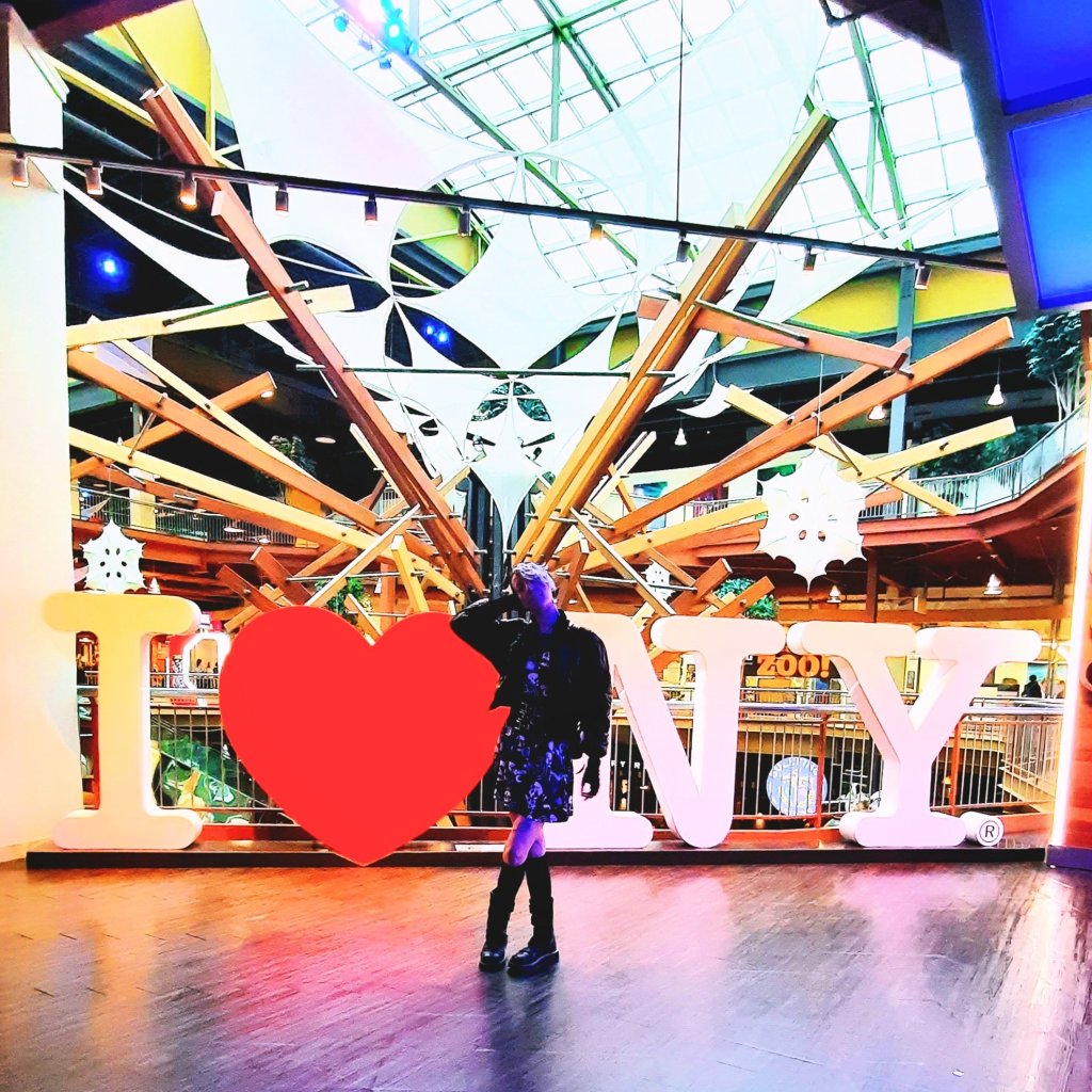 Kae Audhild at Destiny USA mall in Syracuse, NY, Central New York attractions, alternative goth punk lifestyle fashion beauty travel blogger, keracolor silver blue toning semipermanent hair toning color conditioner, killstar x rob zombie official print bardeau style goth punk skater dress, shein black faux leather studded buckle corset style waist belt, shein sxy shiny black puffer bomber jacket, black knee high socks from Amazon, black vegan leather Demonia Lilith-211 mid-calf buckle side zip boots, best of winter 2023 goth fashion, tattooed pierced alt models, thick coarse curly bleached icy blue hair, real natural scandi curls waves, women's afab nonbinary asymmetrical undercut hairstyles