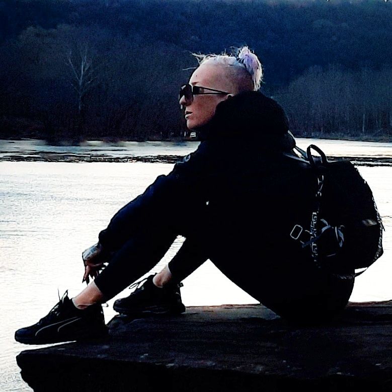 Kae Audhild sitting on a rock by the water in Owego ny, wearing puma twitch trail runners sneakers, shein black mini backpack purse and designer fashion sunglasses, Prozis leggings
