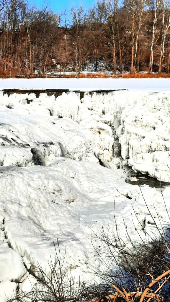 The Beebe Lake dam is iced over, and only a few streams of water are visible. The water continues to run under the ice.