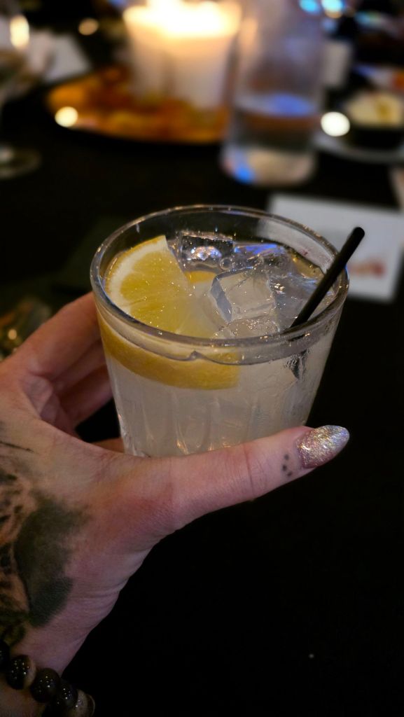 Kae Audhild's hand with holographic glitter short almond gel nails and tattoos holding a cocktail with a lemon wedge, at a fancy networking dinner