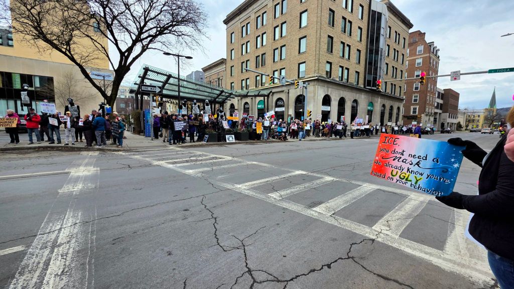 Large crowd gathered at a downtown Ithaca, NY intersection during the ICE Out For Good protest, with demonstrators holding signs and standing in cold, rainy weather.