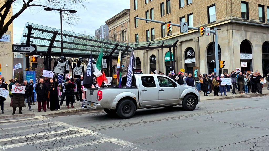 Pickup truck with protest flags driving past a large crowd of demonstrators holding protest signs at the ICE Out For Good rally in downtown Ithaca, New York.