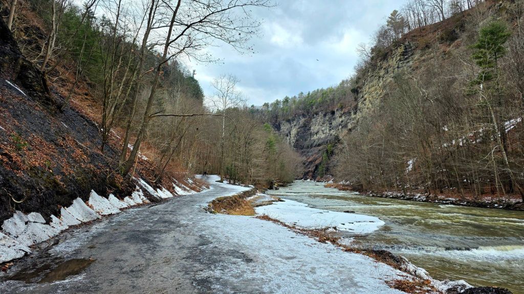 Icy gorge trail at Taughannock Falls State Park in winter, with frozen patches on the path, steep rock walls, and Taughannock Creek flowing beside the trail in the Finger Lakes region of New York.