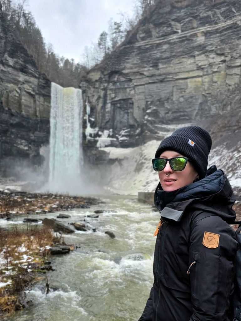 Kae Audhild standing near the base of Taughannock Falls in winter, wearing cold-weather gear with the waterfall and icy gorge walls behind them in Taughannock Falls State Park, New York.