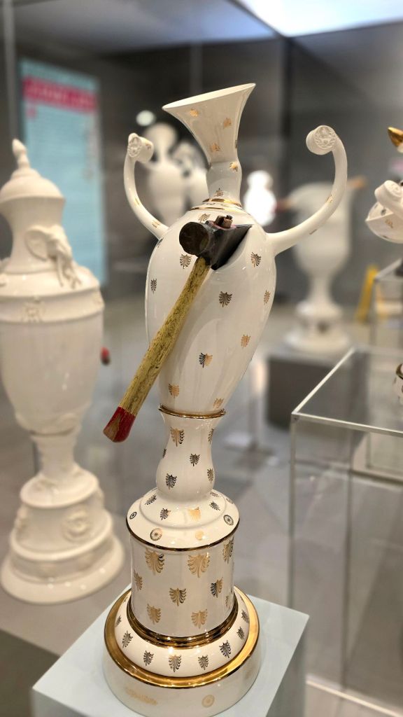 Decorative white ceramic sculpture with gold accents and an axe inserted through the vessel, exhibited in a gallery at the Everson Museum of Art in Syracuse, NY.