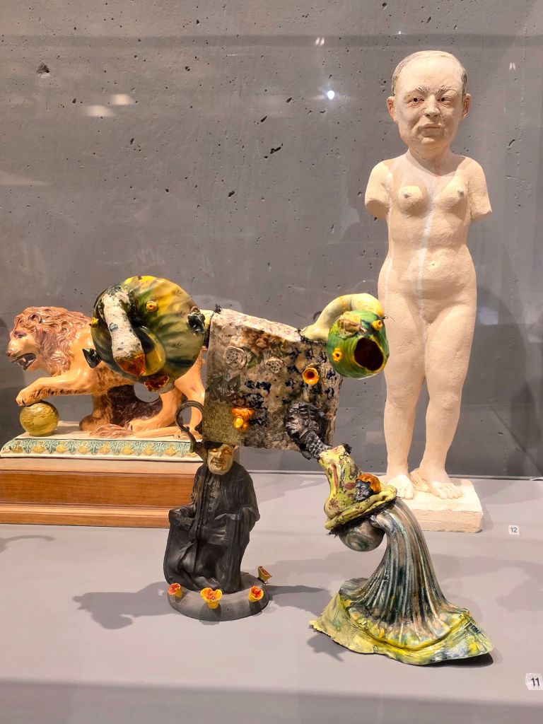 Ceramic figurine sculptures arranged in a museum display, including a distorted animal-like form with textured glaze, a small robed human figure, a reclining lion sculpture, and a pale nude human figure standing in the background, exhibited at the Everson Museum of Art in Syracuse, New York.