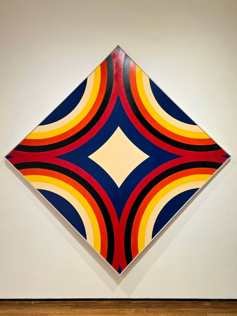 Large diamond-shaped geometric artwork featuring bold curved bands of red, yellow, black, and blue arranged symmetrically on a white gallery wall at the Everson Museum of Art in Syracuse, New York.