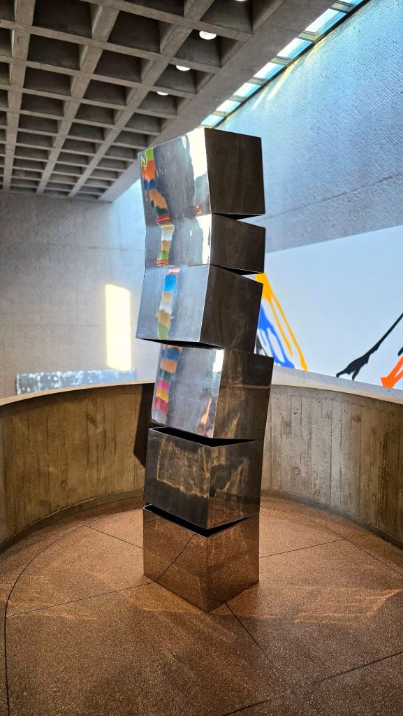Stacked metal geometric sculpture composed of angled, reflective rectangular forms, installed in a concrete gallery space at the Everson Museum of Art in Syracuse, New York.