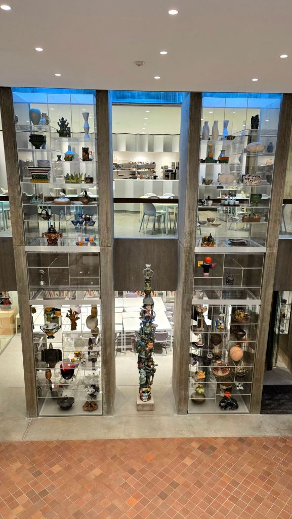 View of tall glass display cases filled with ceramic vessels and sculptural works inside the Everson Museum of Art, with a visible study gallery and seating area in the background in Syracuse, New York.