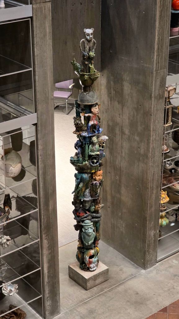 Tall totem-style ceramic sculpture composed of stacked human and animal figures with varied glazes, installed between concrete walls in the Everson Museum of Art in Syracuse, New York.