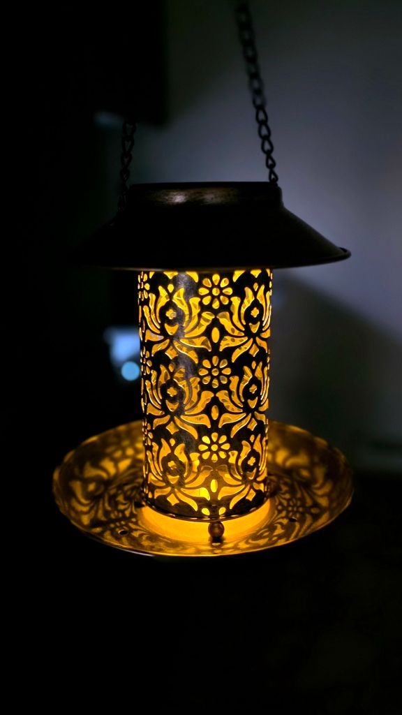 Solar hanging metal bird feeder glowing at night with warm light shining through lotus flower cutout pattern
