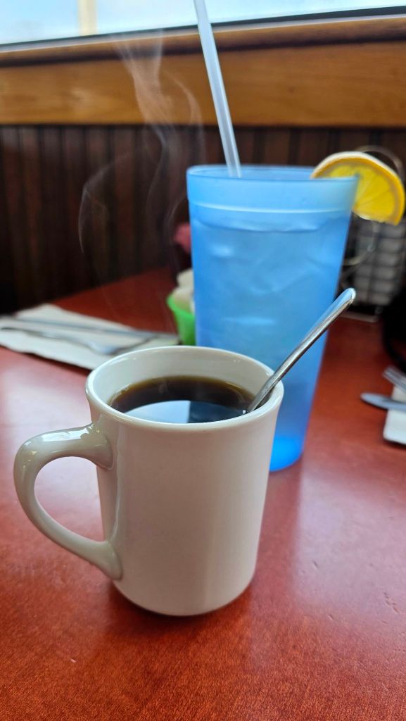 Steaming mug of black coffee with spoon and blue water cup with lemon slice on a diner table at Piccolo’s Cafe in Binghamton, New York.