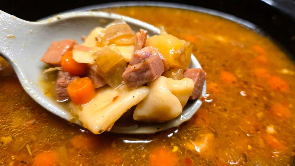 A spoon holding ham pot pie stew with chunks of ham, golden potatoes, carrots, onions, and rustic flat dumplings in a rich broth.