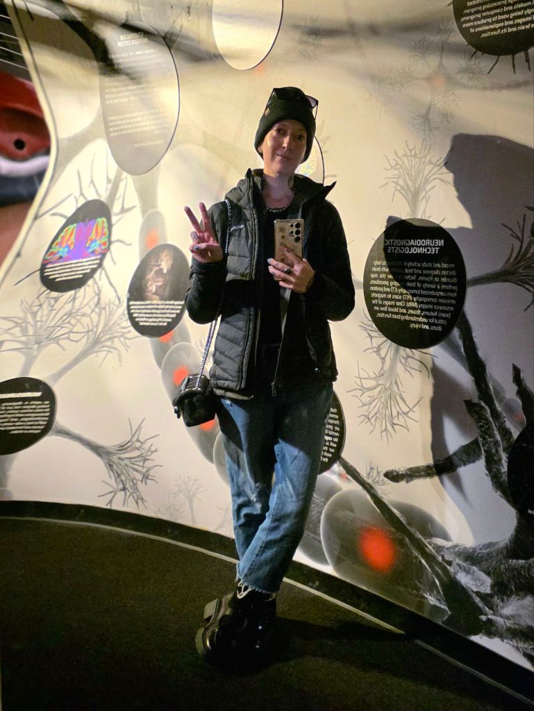 Kae Audhild taking a mirror selfie inside the Museum of Science & Technology in Syracuse, New York, wearing a black beanie, puffer jacket, jeans, and loafers during a winter museum visit.