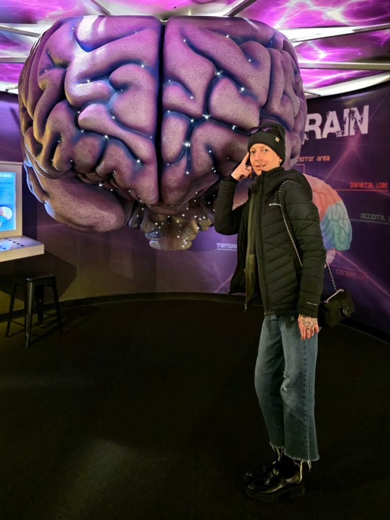 Kae Audhild standing beside a large illuminated brain exhibit at the Museum of Science & Technology in Syracuse, New York, reflecting on mental health, self-care, and emotional well-being during a winter museum visit.