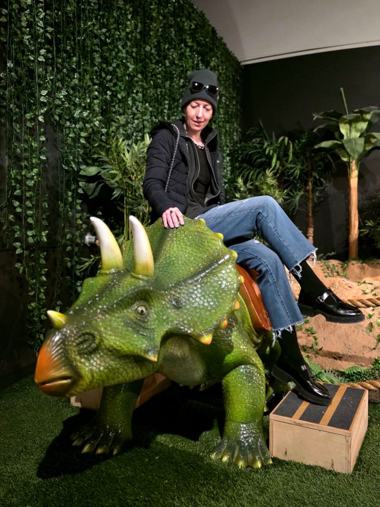 Kae Audhild sitting on a life-size triceratops model inside the dinosaur exhibit at the Museum of Science & Technology in Syracuse, New York during a winter visit.