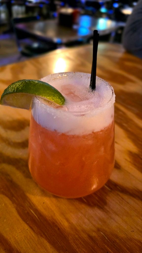 A close-up of the Part Time Lover strawberry margarita at Garage Taco Bar in Binghamton, NY, served in a short glass with a lime wedge garnish and light foam on top, photographed on a warm wood table with soft blue restaurant lighting in the background.