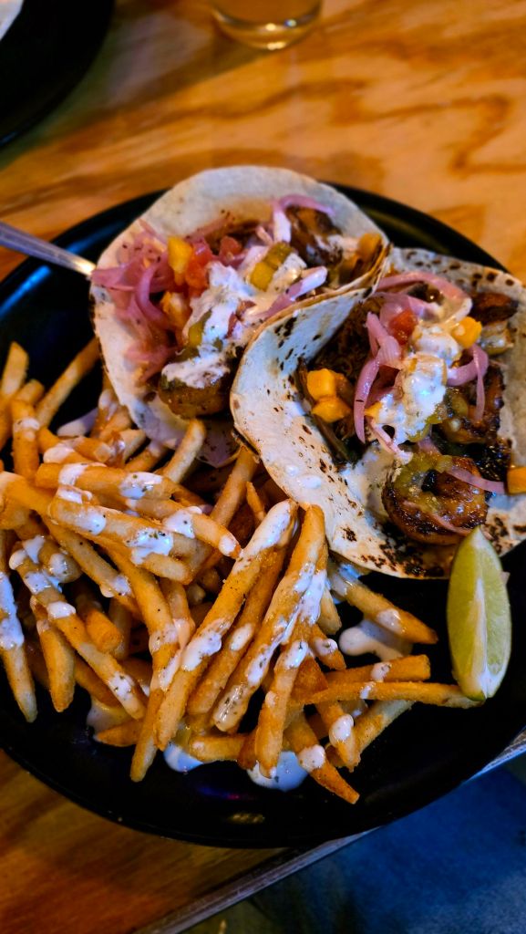 A plate of surf n turf tacos at Garage Taco Bar in Binghamton, NY, topped with mango pico, pickled red onions, and lime crema, served alongside crispy fries drizzled with sauce and a fresh lime wedge.